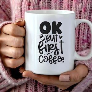 Könnte beinhalten: Eine weiße Keramik-Kaffeetasse mit dem Text "OK but first coffee" in Schwarz. Die Tasse hat einen großen Henkel und wird von einer Person gehalten, die einen rosa Pullover trägt. Die Tasse hat eine Kaffeetassen-Grafik.