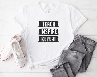 May include: White t-shirt with the words "TEACH INSPIRE REPEAT" in black block letters. The shirt is paired with grey jeans and white trainers with red trim. The items are arranged on a white wooden surface.