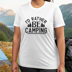 Peut inclure: T-shirt blanc à manches courtes avec l'inscription noire "I'D RATHER BE CAMPING". Le motif comprend deux pins stylisés. Le t-shirt est en matière légère, idéal pour le camping et les activités de plein air.