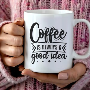 Puede incluir: Una taza de café de cerámica blanca con la inscripción negra "Coffee is always a good idea". La taza tiene un asa grande y la sostiene una persona que lleva un jersey rosa.