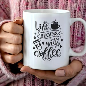 Könnte beinhalten: Weiße Keramik-Kaffeetasse mit dem Schriftzug "Life begins with coffee" in schwarzer Schrift. Die Tasse zeigt kaffeebezogene Grafiken, darunter eine Kaffeetasse und einen To-Go-Becher. Die Tasse wird von einer Person gehalten, die einen rosa Pullover trägt.