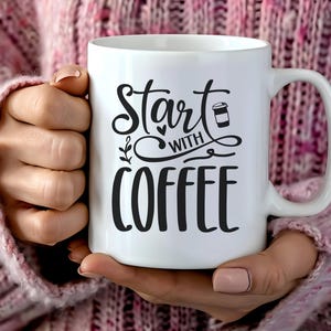 Könnte beinhalten: Weiße Keramik-Kaffeetasse mit dem schwarzen Schriftzug "Start with Coffee". Die Tasse hat einen großen Henkel und wird von einer Person gehalten, die einen rosa Pullover trägt. Die Tasse hat eine Kaffeetassen-Grafik.