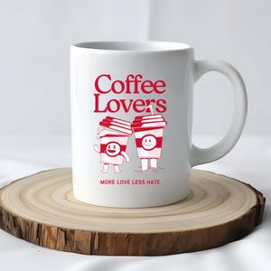 Könnte beinhalten: Weiße Keramik-Tasse mit den roten Worten "Coffee Lovers" und Cartoon-Kaffeetassen-Figuren. Auf der Tasse steht auch der rote Satz "MORE LOVE LESS HATE". Die Tasse steht auf einer Holzscheibe.