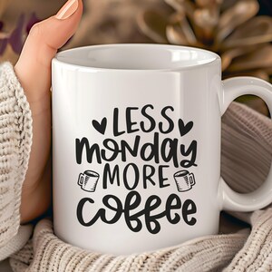 Könnte beinhalten: Weiße Keramik-Kaffeetasse mit dem Text "LESS monday MORE coffee" in schwarzer Schrift. Die Tasse hat einen gebogenen Henkel und wird von einer Person gehalten, die einen cremefarbenen Pullover trägt. Zwei kleine Kaffeetassen-Illustrationen sind enthalten.