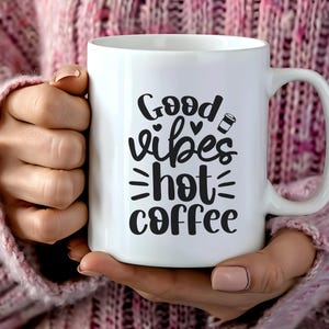 Könnte beinhalten: Weißer Keramik-Kaffeebecher mit den Worten "Good Vibes Hot Coffee" in schwarzer Schrift. Der Becher wird von einer Person gehalten, die einen rosa Pullover trägt. Der Becher hat einen Henkel und ein Kaffeetassen-Symbol.