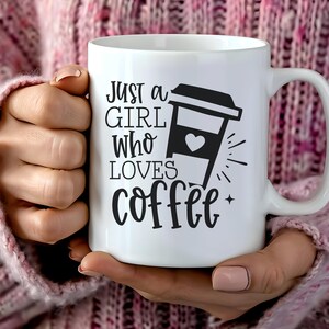 Könnte beinhalten: Weiße Keramik-Kaffeetasse mit dem schwarzen Text "Just a Girl Who Loves Coffee". Die Tasse zeigt eine schwarze Grafik einer Kaffeetasse mit einem Herz. Die Tasse wird von einer Person gehalten, die einen rosa Pullover trägt.
