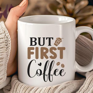 Könnte beinhalten: Weiße Keramik-Kaffeetasse mit dem Text "BUT FIRST Coffee" in Schwarz und Braun. Die Tasse zeigt eine Kaffeetassen-Grafik und Kaffeebohnen-Illustrationen. Die Tasse hat einen gebogenen Henkel.