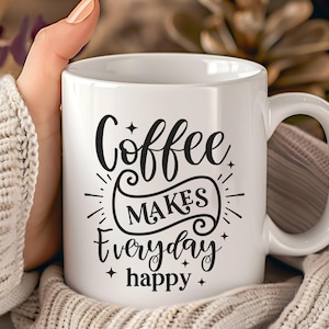 Könnte beinhalten: Weiße Keramik-Kaffeetasse mit dem Text "Coffee makes everyday happy" in schwarzer Schrift. Die Tasse hat einen gebogenen Henkel und wird von einer Person gehalten, die einen cremefarbenen Pullover trägt. Der Hintergrund ist unscharf.