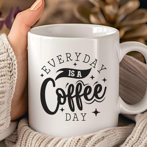 Könnte beinhalten: Weiße Keramik-Kaffeetasse mit dem Text "EVERYDAY IS A Coffee DAY" in schwarzer Schrift. Die Tasse hat einen gebogenen Henkel und wird von einer Person gehalten, die einen cremefarbenen Pullover trägt.
