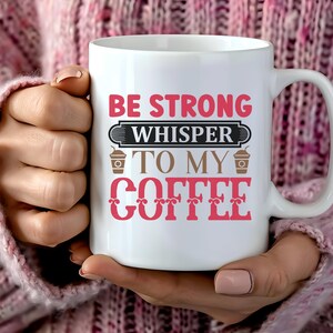 Könnte beinhalten: Weiße Keramik-Kaffeetasse mit dem Text "BE STRONG WHISPER TO MY COFFEE" in Rot und Braun. Die Tasse hat einen großen Henkel und wird von einer Person gehalten, die einen rosa Pullover trägt.