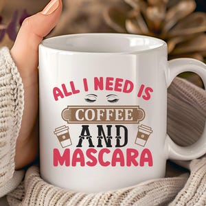 May include: White ceramic coffee mug with the text "ALL I NEED IS COFFEE AND MASCARA" in pink and black. The mug has a handle and two coffee cup graphics. The mug is held by a person wearing a white sweater.