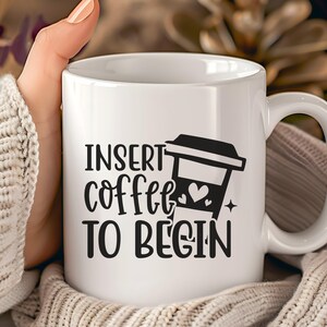 Könnte beinhalten: Weiße Keramik-Kaffeetasse mit dem Text "INSERT coffee TO BEGIN" in Schwarz. Eine schwarze Grafik einer Kaffeetasse mit Herz und Stern befindet sich ebenfalls auf der Tasse. Die Tasse wird von einer Person gehalten, die einen cremefarbenen Pullover trägt.