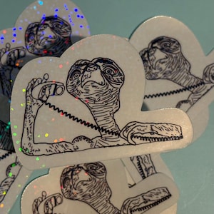 May include: A collection of white, cloud-shaped stickers featuring a black and white illustration of E.T. holding a telephone receiver. The stickers have a holographic, iridescent finish, creating a shimmering effect. The design is a nod to the classic movie.