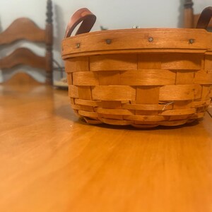 May include: A round, woven wooden basket with a light brown finish, featuring a reinforced rim secured with small metal nails and two arched handles attached to the sides. The basket has a classic splint-weave construction with a flat base.