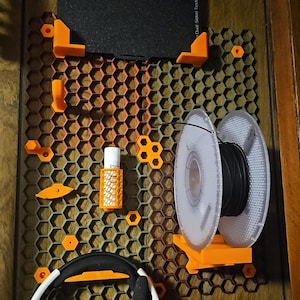 May include: A wall-mounted 3D printing accessory organizer in orange and brown. It features a honeycomb design with various holders for filament spools, tools, and a black print bed. A pair of white and black headphones hangs below.