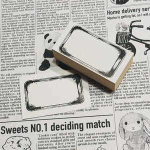 May include: Two rectangular rubber stamps with a decorative border are displayed on a newspaper background. The stamps have a blank white space for text or designs. The image has a vintage aesthetic.