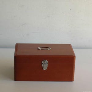 May include: A rectangular wooden storage box with a smooth, medium-brown finish. The box features a silver-coloured metal handle centred on the top lid and a silver-coloured metal latch mechanism on the front panel to secure the lid closed.