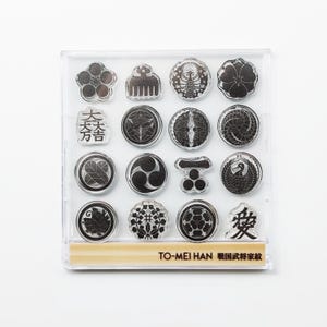 Puede incluir: Una caja de plástico transparente que contiene una colección de sellos de crestas japonesas en blanco y negro. Los sellos presentan varios diseños circulares y florales, junto con caracteres japoneses. El texto "TO-MEI HAN" es visible en la parte inferior.