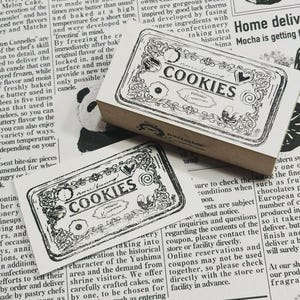 May include: Two rectangular cookie stamps with the word "COOKIES" in a decorative font. The stamps are black and white, with ornate borders and illustrations of cookies. The stamps are on a newspaper background.