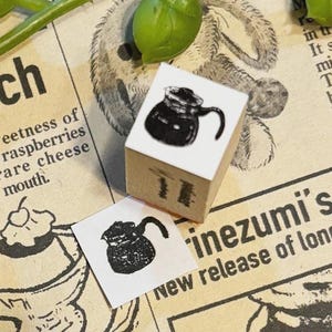 May include: A small, wooden stamp with a black ink illustration of a coffee pot. The stamp is on a newspaper background with the text "ch" and "sweetness of raspberries". A stamped image of the coffee pot is next to the stamp.