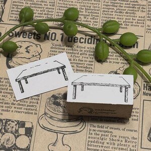 Könnte beinhalten: Zwei Gummistempel mit einem Tischdesign. Ein Stempel befindet sich auf weißem Hintergrund, der andere auf einem braunen Block. Grüne Beeren und Text auf Zeitungshintergrund.