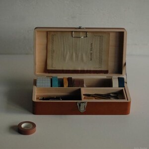 May include: A rectangular wooden storage box with a metal latch and hinged lid. The open lid displays a notebook with the text NOTE BOOK. The interior compartments contain several rolls of patterned washi tape, a black pen, and metal scissors.