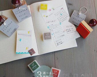 May include: An open notebook with various stationery items. Includes stamps, tags, and stickers with text like "7x7 Planner" and "Message." The color palette is soft, with shades of white, gray, red, and green. The image is taken from an overhead perspective.