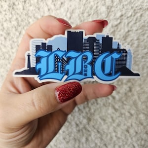 May include: A blue and white sticker with a stylized cityscape and the word "L.B.C." in a bold, gothic font.