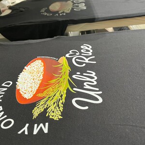 Puede incluir: Una camiseta negra con un estampado gr&aacute;fico de un cuenco de arroz blanco con tallos de grano verdes. El texto "MY ONE AND UNLI RICE" aparece en letras blancas, con un peque&ntilde;o icono de coraz&oacute;n sobre la palabra "Unli" en el centro.