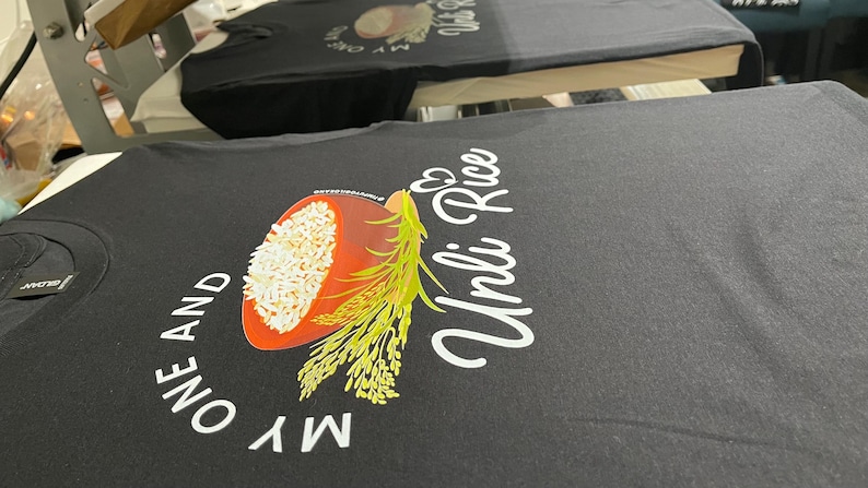 Puede incluir: Una camiseta negra con un estampado gr&aacute;fico de un cuenco de arroz blanco con tallos de grano verdes. El texto "MY ONE AND UNLI RICE" aparece en letras blancas, con un peque&ntilde;o icono de coraz&oacute;n sobre la palabra "Unli" en el centro.