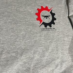 Puede incluir: Una camiseta gris jaspeado con un gr&aacute;fico en el pecho. El dise&ntilde;o muestra un engranaje dividido en mitades roja y negra, con la silueta de un dron negro en el centro. Debajo del engranaje, el texto dice I CAN BECAUSE I AM FORGOTTEN HEROES FOUNDATION.