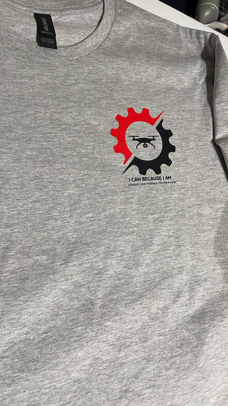 Puede incluir: Una camiseta gris jaspeado con un gr&aacute;fico en el pecho. El dise&ntilde;o muestra un engranaje dividido en mitades roja y negra, con la silueta de un dron negro en el centro. Debajo del engranaje, el texto dice I CAN BECAUSE I AM FORGOTTEN HEROES FOUNDATION.