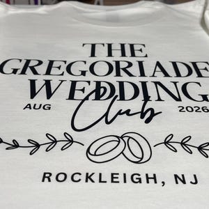 Puede incluir: Una camiseta blanca con texto negro que dice THE GREGORIADES WEDDING CLUB, AUG 2026 y ROCKLEIGH, NJ. El dise&ntilde;o incluye un gr&aacute;fico negro de dos anillos de boda entrelazados centrados entre dos ilustraciones horizontales de ramas con hojas.