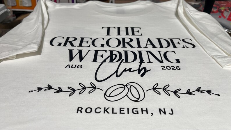 Puede incluir: Una camiseta blanca con texto negro que dice THE GREGORIADES WEDDING CLUB, AUG 2026 y ROCKLEIGH, NJ. El dise&ntilde;o incluye un gr&aacute;fico negro de dos anillos de boda entrelazados centrados entre dos ilustraciones horizontales de ramas con hojas.