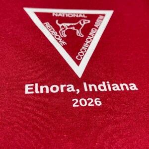 Puede incluir: Una camiseta de color rojo oscuro con un gr&aacute;fico de tri&aacute;ngulo blanco que contiene el texto NATIONAL REDBONE COONHOUND ASS'N y la silueta de un perro. Debajo del tri&aacute;ngulo, el texto Elnora, Indiana 2026 est&aacute; impreso en letras blancas.