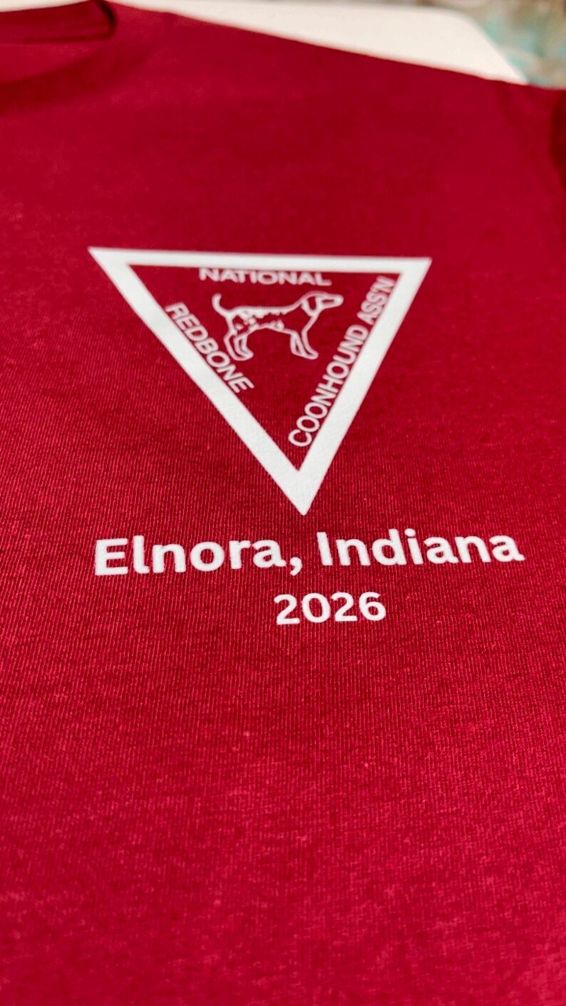 Puede incluir: Una camiseta de color rojo oscuro con un gr&aacute;fico de tri&aacute;ngulo blanco que contiene el texto NATIONAL REDBONE COONHOUND ASS'N y la silueta de un perro. Debajo del tri&aacute;ngulo, el texto Elnora, Indiana 2026 est&aacute; impreso en letras blancas.