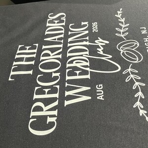 Puede incluir: Una camiseta negra con texto impreso en blanco que dice THE GREGORIADES WEDDING CLUB AUG 2026, acompa&ntilde;ada de un gr&aacute;fico de dos anillos entrelazados y una rama con hojas, con la ubicaci&oacute;n ROCKLEIGH, NJ impresa verticalmente.
