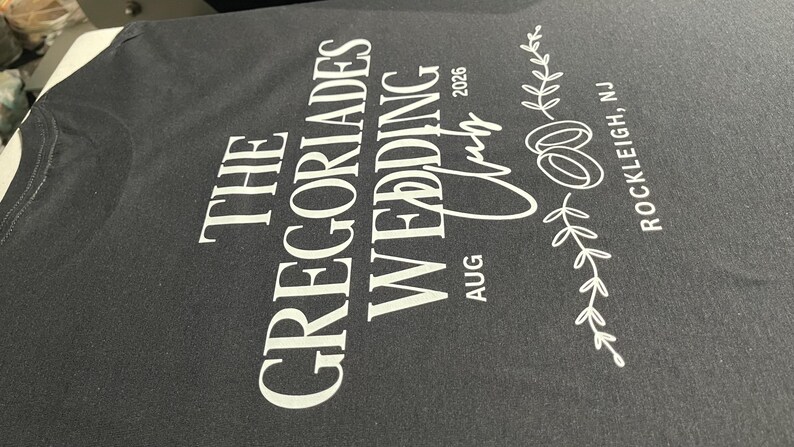 Puede incluir: Una camiseta negra con texto impreso en blanco que dice THE GREGORIADES WEDDING CLUB AUG 2026, acompa&ntilde;ada de un gr&aacute;fico de dos anillos entrelazados y una rama con hojas, con la ubicaci&oacute;n ROCKLEIGH, NJ impresa verticalmente.
