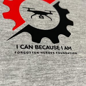 Puede incluir: Una camiseta gris jaspeado con un gr&aacute;fico de un dron negro centrado dentro de un icono de engranaje dividido en secciones roja y negra. Debajo del gr&aacute;fico, el texto dice I CAN BECAUSE I AM y FORGOTTEN HEROES FOUNDATION en color negro.