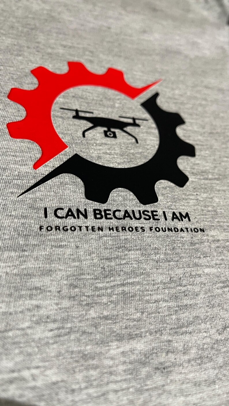 Puede incluir: Una camiseta gris jaspeado con un gr&aacute;fico de un dron negro centrado dentro de un icono de engranaje dividido en secciones roja y negra. Debajo del gr&aacute;fico, el texto dice I CAN BECAUSE I AM y FORGOTTEN HEROES FOUNDATION en color negro.