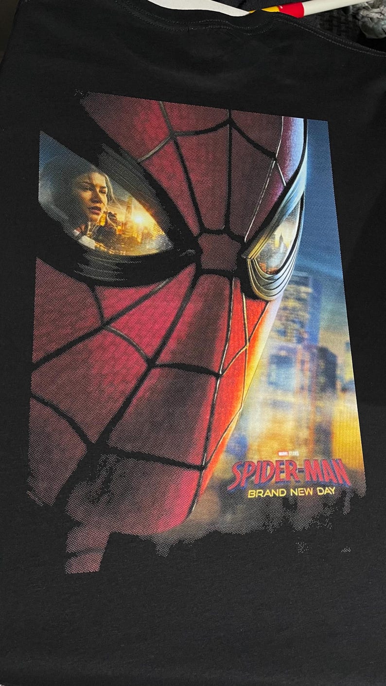 Puede incluir: Una camiseta negra con un gran estampado gr&aacute;fico de la m&aacute;scara de Spider-Man en rojo y negro. Las lentes de la m&aacute;scara reflejan una escena urbana y una persona. El texto "SPIDER-MAN" y "BRAND NEW DAY" aparece en rojo y amarillo abajo.