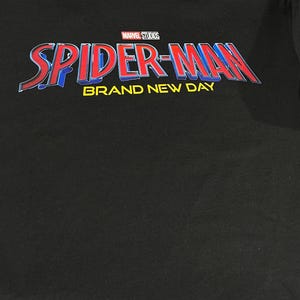 Puede incluir: Una camiseta negra de cuello redondo con el logotipo de Marvel Studios sobre el texto SPIDER-MAN en letras rojas y azules, con las palabras BRAND NEW DAY impresas en amarillo debajo. La camiseta tiene una etiqueta de la marca Gildan en el cuello.