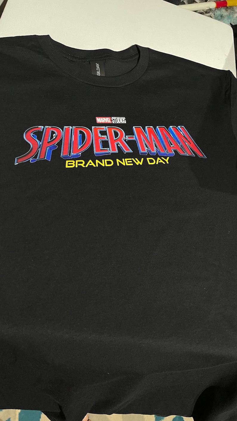 Puede incluir: Una camiseta negra de cuello redondo con el logotipo de Marvel Studios sobre el texto SPIDER-MAN en letras rojas y azules, con las palabras BRAND NEW DAY impresas en amarillo debajo. La camiseta tiene una etiqueta de la marca Gildan en el cuello.