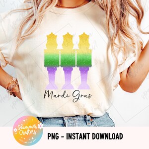 Könnte beinhalten: Cremefarbenes T-Shirt mit einem Mardi Gras-Design. Die Grafik zeigt drei stilisierte Figuren in Gelb, Grün und Lila, mit dem Schriftzug "Mardi Gras" in Schreibschrift. Das Design ist ein PNG-Sofort-Download.