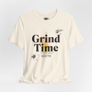 Può includere: T-shirt color crema con le parole "Grind Time" in grandi lettere nere. Sotto il testo c'è "A Summer Collection by LMR Threadz" e "XXIV/VII". La maglietta presenta disegni a spruzzi neri e un gradiente giallo e arancione.