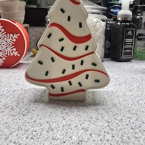 Op de afbeelding: Een crèmekleurige kerstboomvormige decoratie met rode golvende lijnen en kleine groene rechthoeken. De boom heeft een platte basis en staat op een gespikkelde aanrecht. Andere items zijn zichtbaar op de achtergrond.