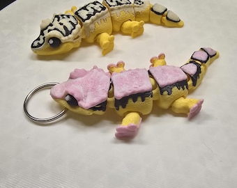 May include: Two small, yellow, segmented gecko figurines. One gecko has black and white markings and the other has pink and black markings. The gecko with pink markings has a silver key ring attached.