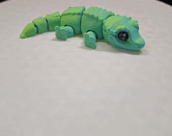 May include: A green and blue segmented gecko toy with black eyes. The toy is made of plastic and has a segmented body.