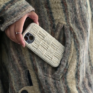 Può includere: Una custodia per telefono color crema con un motivo intrecciato è tenuta in tasca. La custodia ha ritagli per la fotocamera e il flash. Lo sfondo è un cappotto strutturato, multicolore, nei toni del marrone, verde e beige.
