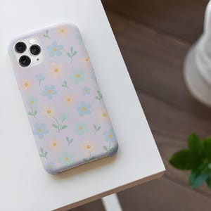 Puede incluir: Una funda de teléfono lavanda con un estampado floral. El diseño presenta pequeñas flores azules, amarillas y verde claro con hojas verdes. La funda está sobre una superficie blanca.